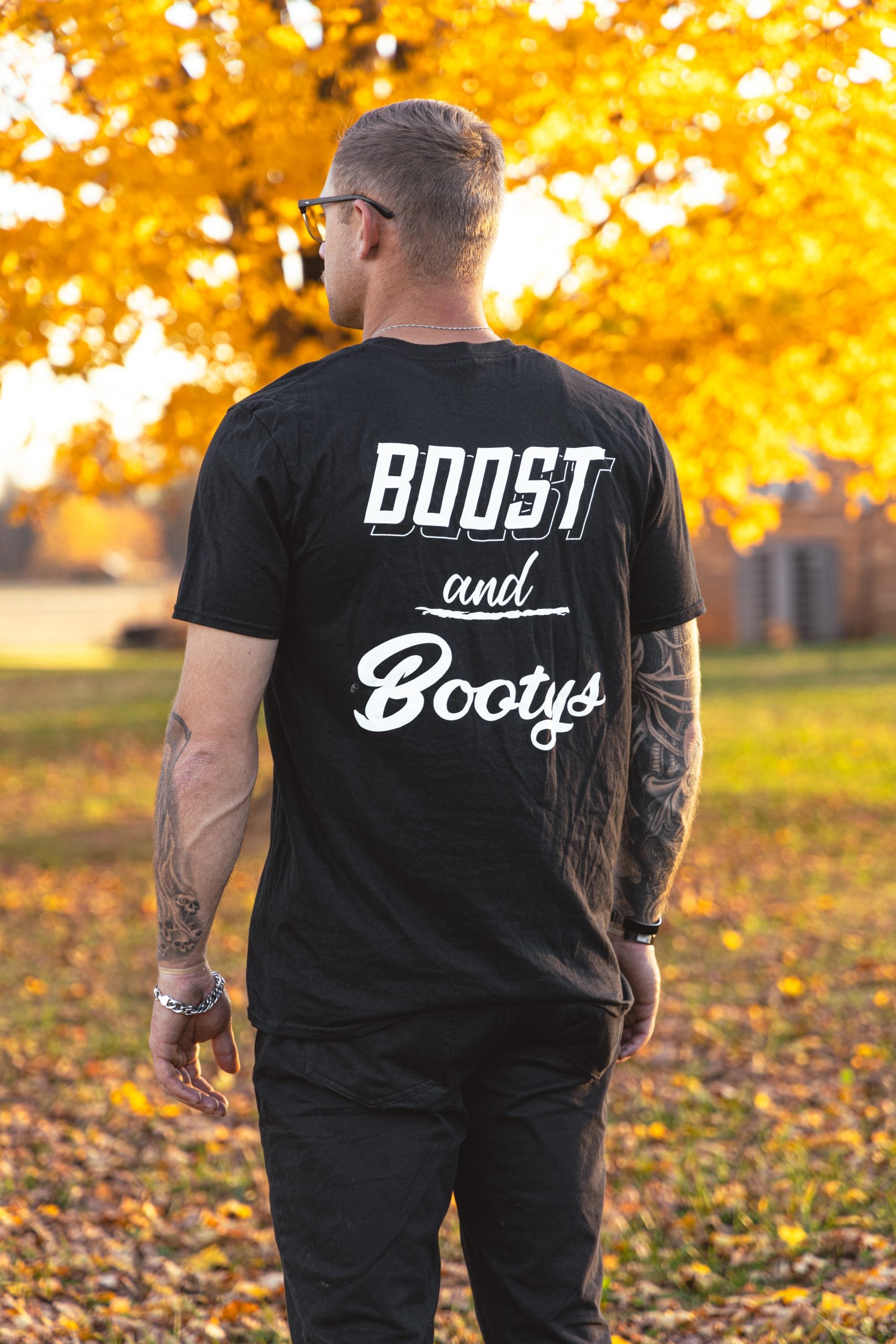 Man wearing a black t-shirt with 'Boost and Bootys' text in an outdoor setting with trees in the background.